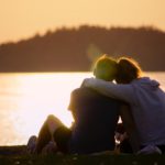 Two people sitting together and hugging while facing a water body