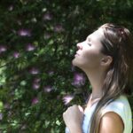 Woman Closing Her Eyes Against Sun Light Standing Near Purple Petaled Flower Plant